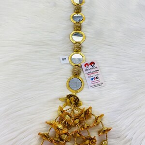 May include: A gold beaded chain with round mirrors and a gold tassel with multiple layers of gold fabric flowers. The chain is hanging on a white fuzzy surface. The number "24" is visible on a white tag.