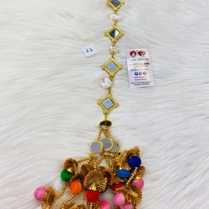 May include: A gold chain with small mirrors and white beads, ending in a tassel with colorful pom-poms and gold accents. The chain has a number "22" on the top left corner. The image also includes a business card with the text "Safa Turban +1-437-635-3040 Follow Us On: Instagram: @safaturban Facebook: @safaturban Shop 5, 108 Kennedy Rd, South Brampton, ON L6W 3P4"