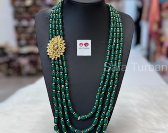 Puede incluir: Un collar de cuentas verdes con un colgante dorado con un diseño floral. El collar está formado por cuatro hileras de cuentas.