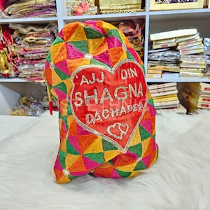 Può includere: Una borsa rossa a forma di cuore con motivi geometrici colorati e testo ricamato che recita "AJJ DIN SHAGNA DACHADER".