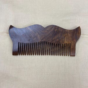 Peut inclure: Un peigne en bois brun foncé avec une finition lisse et polie et un motif de grain distinct. Le peigne présente un bord supérieur large et incurvé qui se termine par deux extrémités pointues, avec une série de dents fines et régulières.
