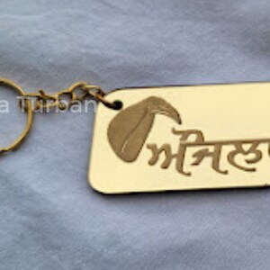 May include: Gold-colored keychain with a small gold chain and a rectangular tag. The tag has a gold-colored image of a turban and the text "Safa Turban" and the word "ਮੀਂਹੁ" in Punjabi.