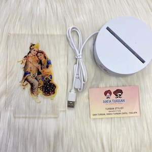 May include: A white acrylic light-up base with a clear acrylic insert featuring a colorful image of a couple. The base is connected to a USB cable and a small switch. The packaging includes a card with the text "SAFA TURBAN TURBAN STYLIST Specialist in Tying SIKH TURBAN, HINDU TURBAN (SAFA), THALAPA".