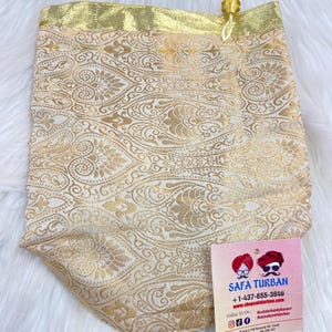Puede incluir: Una bolsa de cordón de color dorado con un patrón de cachemira. La bolsa está hecha de un tejido suave y sedoso y tiene un borla dorada. La bolsa está sentada sobre una superficie blanca. La bolsa tiene una tarjeta de visita adjunta con el texto "Safa Turban" e información de contacto.