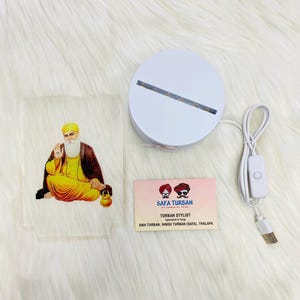 Peut inclure: Une base de lumière LED ronde blanche avec un panneau acrylique transparent représentant une illustration colorée d'un homme sikh portant un turban. La base est équipée d'un câble USB et d'un interrupteur marche/arrêt. Une carte de visite avec le texte "SAFA TURBAN TURBAN STYLIST Spécialiste du nouage SIKH TURBAN, HINDU TURBAN (SAFA), THALAPA" est également incluse.