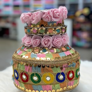 May include: A traditional Indian ceremonial pot decorated with pink fabric flowers, gold sequins, colorful tassels, and mirrored accents.