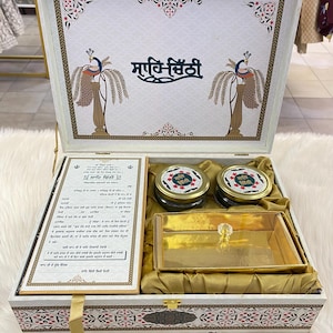 Peut inclure: Coffret cadeau en bois blanc orné de motifs floraux et de paons. À l'intérieur, un plateau doré, deux pots et une carte imprimée. La boîte est doublée de tissu doré et porte le texte "ਸਾਹਿਬਿ ਬਿਂਦੀ" sur le couvercle.