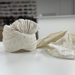 May include: An ivory-coloured turban with detailed embroidered patterns. A matching scarf is draped alongside. The fabric has a textured appearance, possibly woven. The turban is likely for formal events, with a traditional design.