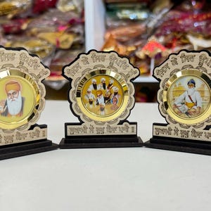 May include: Three small wooden plaques with gold accents and images of Sikh religious figures. The plaques have the text "Guru Nanak Dev Ji" written on them.