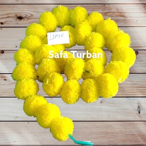 Puede incluir: Una larga guirnalda de pompones de flores de caléndula de color amarillo brillante enhebrados. La guirnalda está sobre una superficie de madera. El texto "Lemon" y "Safa Turban" es visible en una etiqueta blanca.