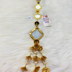 May include: A gold-colored hanging decoration with a large, square mirror at the bottom. The mirror is surrounded by a gold frame and has a white, sparkly surface. The decoration has a chain of smaller, round mirrors above the large mirror, and a series of gold tassels with white beads and red, green, and yellow pom-poms at the bottom.