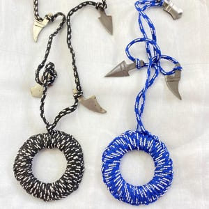Puede incluir: Primer plano de collares hechos a mano. Un collar presenta una cuerda en blanco y negro con un colgante circular y un dije de punta de flecha de metal. Otro collar tiene una cuerda azul y blanca con un colgante circular.