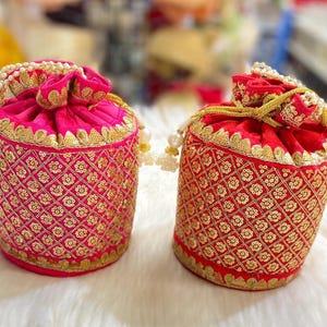 May include: Two ornate, cylindrical drawstring bags. One is hot pink, the other is red. Both are embellished with gold embroidery and sequins in a floral pattern. The tops are gathered with a gold cord and adorned with white pearl-like beads.