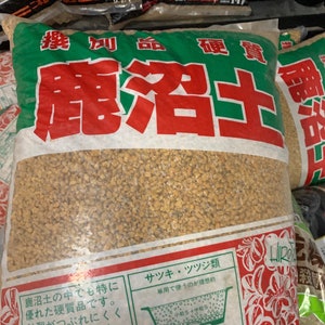 Peut inclure: Un sac de terreau granulaire brun, étiqueté "鹿沼土" (sol de Kanuma) en japonais. Le sac contient des instructions pour l'utilisation du terreau pour diverses plantes, notamment les azalées, les rhododendrons et les plantes de montagne. Le texte mentionne également la taille idéale des particules du terreau pour différents types de plantes.