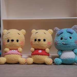 Puede incluir: Tres animales de peluche tejidos a crochet: un oso amarillo con una camisa rosa, un oso amarillo con una camisa roja y un monstruo azul con una camisa blanca.