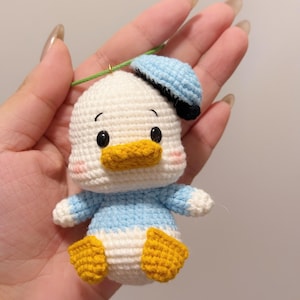 May include: A crocheted duck plush toy with a blue hat. The duck is white with yellow feet and a light blue shirt. The duck has black eyes and a yellow beak.