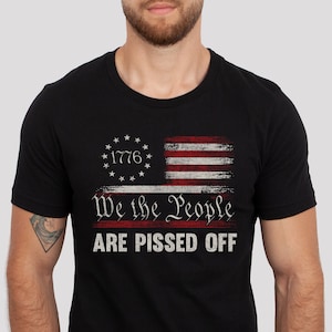 May include: Black t-shirt featuring a distressed American flag design with the text "1776" and "We the People ARE PISSED OFF" in white and red. The shirt has a crew neck and short sleeves.