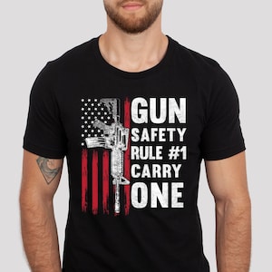 May include: Black crew neck t-shirt with a distressed American flag design and a rifle graphic. White text reads "GUN SAFETY RULE #1 CARRY ONE". The design is printed on the front of the shirt.