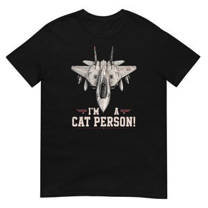 May include: A black short-sleeve t-shirt featuring a graphic of a gray fighter jet centered on the chest. Below the jet, the text reads "I'M A CAT PERSON!" in white capital letters, accented by horizontal red lines and small stars on each side.