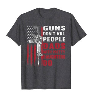 May include: Dark gray t-shirt featuring a distressed American flag design with a rifle graphic. The text reads "GUNS DON'T KILL PEOPLE DADS WITH PRETTY DAUGHTERS DO" in white and red.