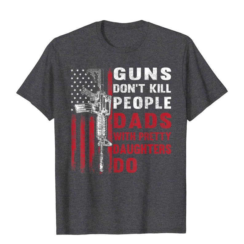 May include: Dark gray t-shirt featuring a distressed American flag design with a rifle graphic. The text reads "GUNS DON'T KILL PEOPLE DADS WITH PRETTY DAUGHTERS DO" in white and red.
