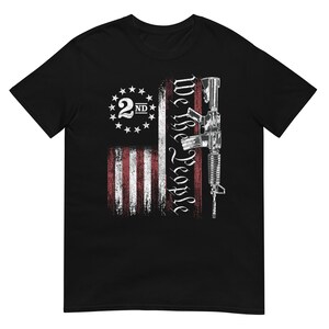 Puede incluir: Camiseta negra con un diseño desgastado de la bandera estadounidense, el texto "We The People" y un gráfico de rifle. El diseño incluye un símbolo de la "2ND" enmienda.