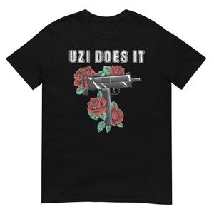 May include: Black t-shirt with the text "UZI DOES IT" in white block letters. A graphic of a silver handgun is surrounded by red roses with green leaves. The shirt is made of a soft material.
