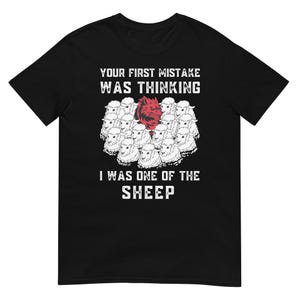 Peut inclure: T-shirt noir avec le texte "YOUR FIRST MISTAKE WAS THINKING I WAS ONE OF THE SHEEP." Le graphisme représente un groupe de moutons blancs avec un loup rouge au centre.