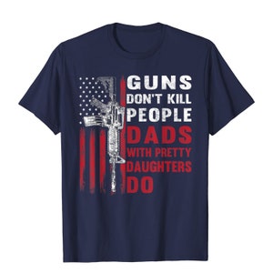 May include: Navy blue t-shirt featuring a distressed American flag design with a rifle graphic. The text reads "GUNS DON'T KILL PEOPLE DADS WITH PRETTY DAUGHTERS DO" in white and red.