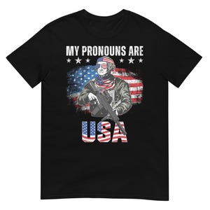 May include: Black t-shirt with a graphic featuring a patriotic design. The design includes an illustration of a person in sunglasses and a tricolor scarf, holding a rifle in front of an American flag. The text "MY PRONOUNS ARE" and "USA" are also displayed.