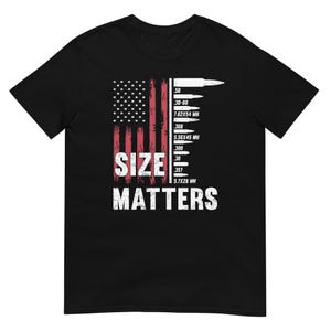 Può includere: T-shirt nera con un design a bandiera americana invecchiata e la scritta bianca "SIZE MATTERS". Il design include una tabella delle dimensioni dei proiettili, con misure in millimetri.