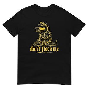 May include: A black short-sleeve t-shirt featuring a yellow graphic of a snake coiled around a security camera. Below the illustration, the text reads "don't flock me" in a stylized yellow font. The design includes small patches of grass.