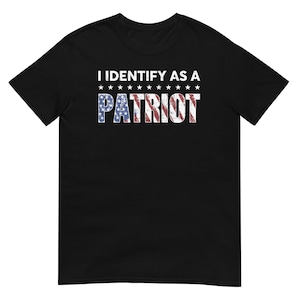 Puede incluir: Camiseta negra con el texto blanco "I IDENTIFY AS A PATRIOT". La palabra "PATRIOT" está rellena con un diseño de bandera americana desgastada, con estrellas blancas encima. Prenda de vestir informal.