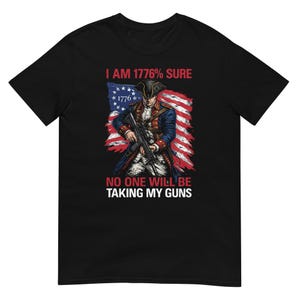 Peut inclure: T-shirt noir avec un motif patriotique. Le graphisme comprend une illustration d'un homme en uniforme de la guerre d'Indépendance tenant un fusil, sur fond de drapeau américain stylisé. Le texte dit "I AM 1776% SURE" et "NO ONE WILL BE TAKING MY GUNS."