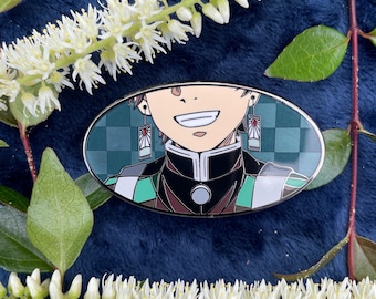 May include: A green, black, and white enamel pin featuring a character from the anime Demon Slayer. The character is smiling and has red earrings with a white sun design. The background is a green and white checkered pattern.