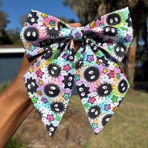 May include: A fabric hair bow featuring a repeating pattern of black soot sprites with large white eyes, interspersed with colorful stars against a pastel rainbow checkered background. The bow has two long, pointed tails and a central fabric knot.