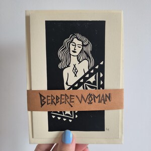 Peut inclure: Un lot de cartes de couleur crème avec une impression linogravure en noir et blanc d'une femme. L'impression est enveloppée d'une bande de papier marron avec l'inscription "BERBERE WOMAN" en lettres noires. La femme a de longs cheveux ondulés.