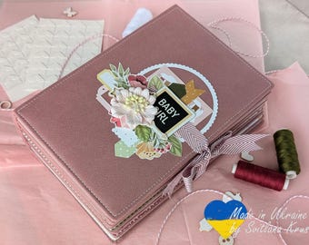 May include: A dusty rose scrapbook album with floral embellishments and the words "Baby Girl" on a small chalkboard. The album is tied with a striped ribbon and sits on pink tissue paper, with spools of thread nearby.