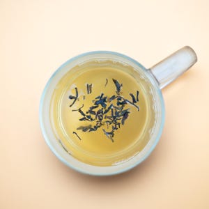 May include: A clear glass mug with a handle filled with light yellow tea and loose tea leaves.