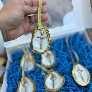 May include: Handmade oyster shell ornaments with gold trim and a gold cross. Each ornament has a gold ribbon for hanging. The ornaments are displayed in a white box with blue paper filler.