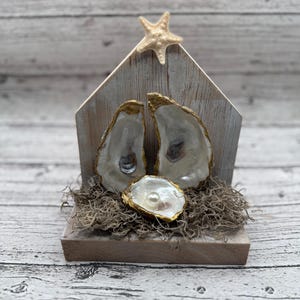 May include: A rustic wooden nativity scene featuring oyster shells with gold trim and a pearl. A small starfish adorns the top of the wooden structure, and the base is covered in dried moss.