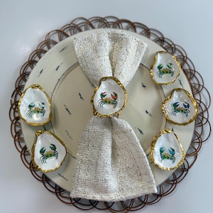 May include: A set of six oyster shell dishes and one matching napkin ring, each featuring a hand-painted blue and orange crab design on a white interior with a metallic gold-painted rim. The napkin ring holds a textured, off-white fabric napkin.