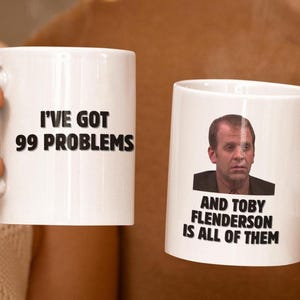 以下が含まれることがあります： 2つの白いセラミックマグ。1つのマグには黒字で「I'VE GOT 99 PROBLEMS」と書かれています。もう1つのマグには男性の写真と黒字で「AND TOBY FLENDERSON IS ALL OF THEM」と書かれています。