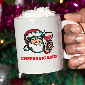 May include: White ceramic mug with a cartoon Santa Claus design, holding a glass of red wine. The Santa illustration has a red hat and white beard. The mug has the text "CHEERS BIG EARS" in red. The mug is filled with whipped cream.
