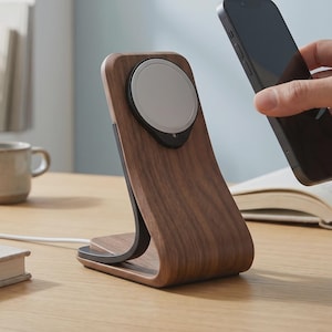 Può includere: Un supporto di ricarica wireless in legno scuro con una base di ricarica magnetica circolare e un cavo di alimentazione bianco. Il supporto presenta un design curvo e verticale con una struttura di supporto interna nera.