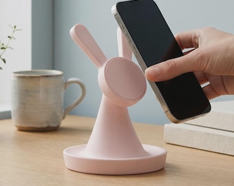 May include: A light pink wireless charging stand shaped like a rabbit with two upright ears and a circular magnetic charging pad. The stand features a wide, circular base and is designed to hold a black smartphone in a vertical position.