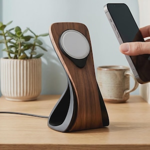 Può includere: Un supporto di ricarica wireless con un pannello frontale in legno scuro e una base nera, dotato di una piastra di ricarica magnetica bianca circolare integrata nella parte superiore, collegata da un sottile cavo di alimentazione nero.
