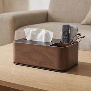 Puede incluir: Un soporte rectangular para caja de pañuelos de madera oscura con un panel superior negro y un compartimento lateral integrado. El compartimento contiene un mando a distancia negro, uno blanco y unas gafas de montura marrón. Sobresalen pañuelos blancos.