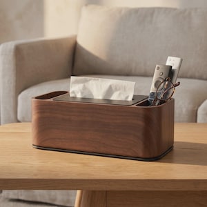 Puede incluir: Un soporte rectangular para caja de pañuelos de madera oscura con un panel superior negro que tiene una ranura central para pañuelos y un compartimento lateral para guardar objetos pequeños como mandos a distancia y gafas.