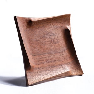 Puede incluir: Una bandeja cuadrada de madera con un acabado de nogal marrón oscuro. La bandeja tiene un diseño ligeramente curvo y ondulado y un borde elevado.